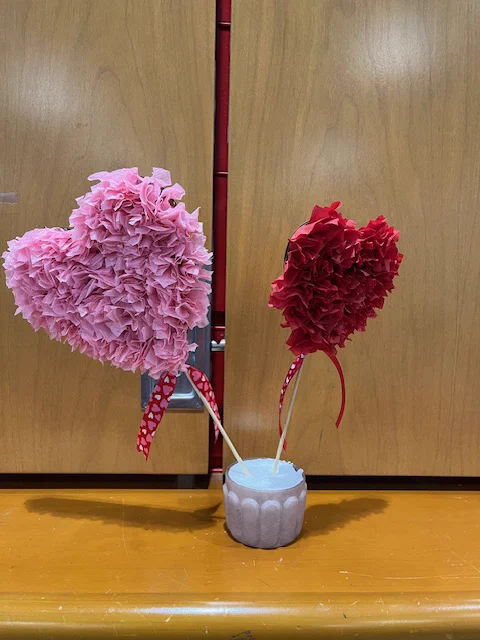 A pink and red hearts filled with rolled up crepe paper florets were placed on sticks and placed upright in a small container.