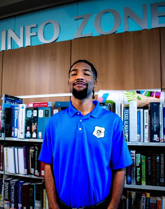 A young man in a blue shirt with a security patch poses in front of a book shelf with a sign that reads "Info Zone" above it.