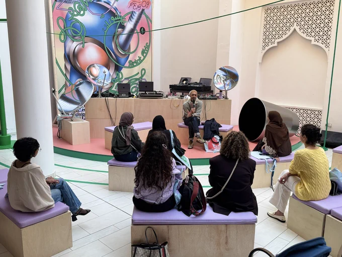 This wide shot captures an outdoor talk or performance in a brightly lit, modern courtyard. Several people are seated on lavender-cushioned wooden benches, facing a speaker.
The Setting
• The Speaker: A person sits on a raised platform, holding a microphone and gesturing as they speak. Behind them is a DJ setup with turntables and laptops.
• Decor: A massive, colorful pop-art mural featuring blue and pink spheres and tangled green lines dominates the back wall. Two large silver satellite dishes are repurposed as decorative elements on the stage.
• Architecture: The space features a white wall with an intricate, geometric latticework archway (mashrabiya style) and a large, black megaphone-shaped sculpture on the floor.
The Audience
The audience, mostly women, is seen from behind. They are dressed casually in sweaters and jeans, listening attentively in the open-air space.