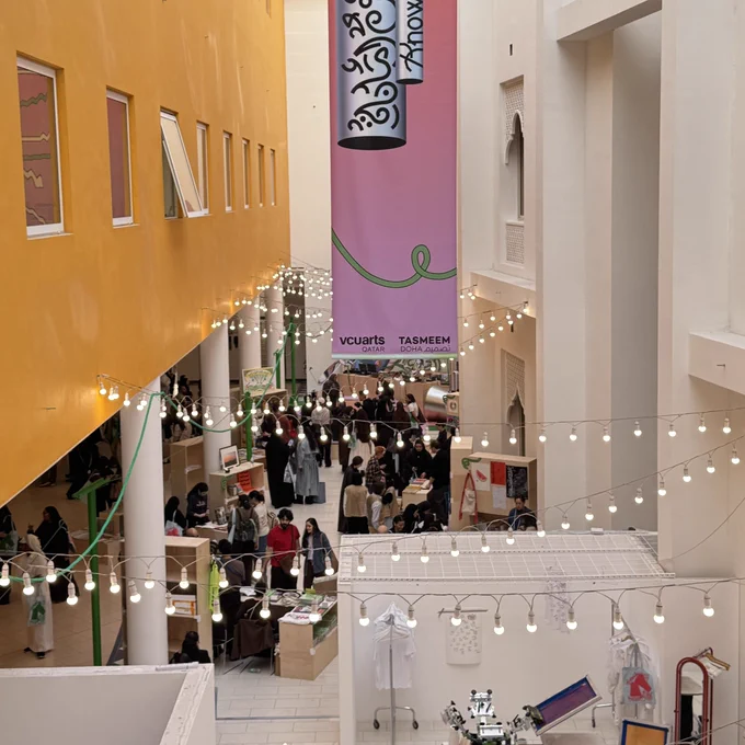 This high-angle shot provides an overview of the Tasmeem Doha art and design biennial at VCUarts Qatar. The scene is a bustling indoor courtyard filled with exhibitors and visitors.
The Venue
• Layout: A narrow, high-ceilinged walkway is flanked by a bright yellow wall with windows on the left and a white wall with arched, traditional-style windows on the right.
• Lighting: Rows of warm, hanging bulb string lights crisscross the space, creating a festive atmosphere.
• Centerpiece: A massive pink vertical banner hangs from the ceiling, featuring a green squiggly line and the "vcuarts QATAR" and "TASMEEM DOHA" logos at the bottom.
Activity
• Marketplace: The ground floor is filled with small wooden stalls and tables where artists display books, prints, and apparel.
• Crowd: Dozens of people are seen browsing the stalls, chatting, and walking through the vibrant, creative marketplace.