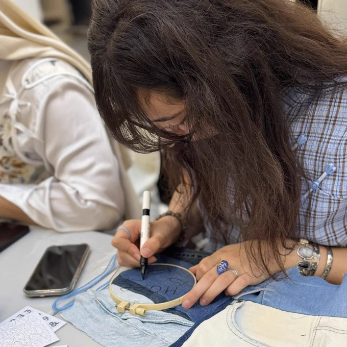 This image captures a close-up, high-angle view of a person engaged in a detailed embroidery and textile project. The focus is on their hands and the tools they are using at a white table.
The Crafting Process
• Central Action: A person with long, dark wavy hair leans over a small wooden embroidery hoop. They are using a black fabric marker to write the word "HOPE" onto a piece of dark blue denim stretched within the hoop.
• Materials: Around the hoop are various scraps of light and dark blue denim. A strand of bright blue yarn and a sheet of letter stickers are also visible on the table.
• Personal Style: The crafter wears several silver rings, including one with a large blue stone, and a silver watch. They are dressed in a blue and white checkered shirt.
The Environment
In the background, another person wearing a cream-colored headscarf and a patterned vest is partially visible, also working at the table. A smartphone sits nearby on the white surface