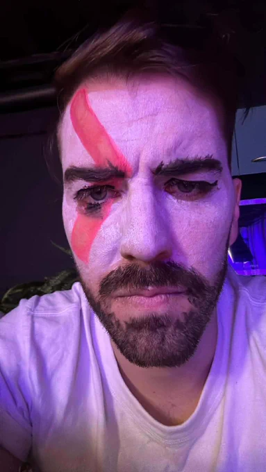 Zac is frowning at the camera with his face painted white and a red stripe on his eye to resemble Kratos, from the God of War games.