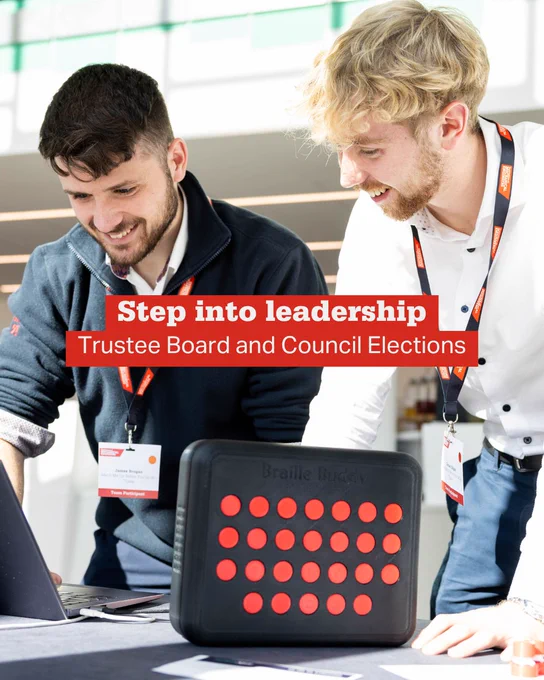 Two men wearing conference badges engage with a Braille Buddy device on a table during a leadership event.