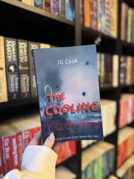 A hand in a cream long sleeve holds a paperback book titled The Cooling by JG Cook. The cover features a dark, moody blue design with red title text and subtle abstract details. The tagline near the bottom reads that winning is easy if you ignore the rules. Behind the book is a softly blurred bookshelf filled with neatly arranged, colourful books.