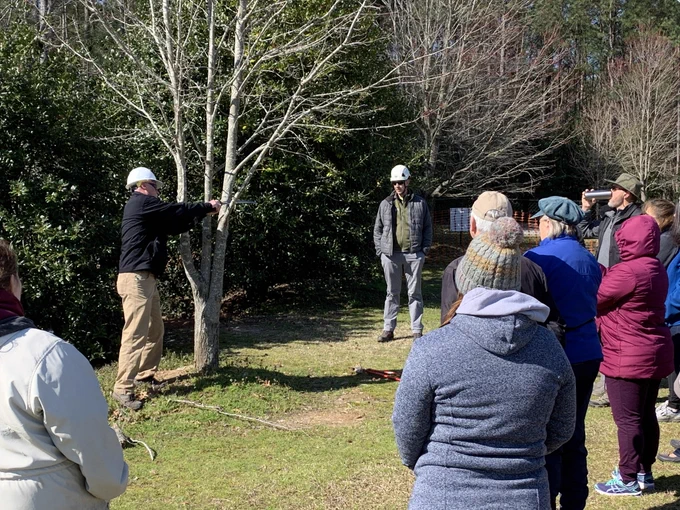 A group of people stands outside, observing a person demonstrating near a tree. Several individuals are wearing helmets, including the demonstrator who is pointing towards the tree. The setting is a grassy area with other trees and shrubs in the background, suggesting a workshop or outdoor educational event.