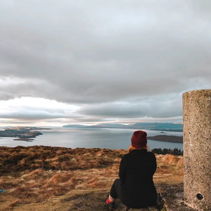Kirsti facing away from the camera sitting at the top of a hill looking out to the sea