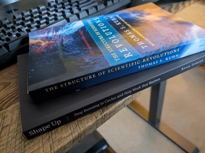 Two books stacked in front of a keyboard. 'The Structure of Scientific Revolutions' by Thomas Kuhn, and 'Shape Up' by Ryan Singer 