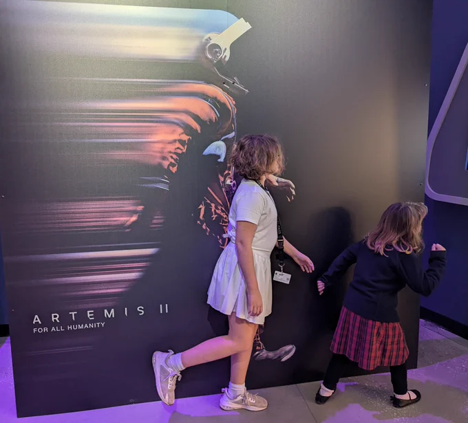 Children walking in front of a mural of an astronaut walking in the Artemis II space suit. The children are wearing skirts and have short light brown hair.