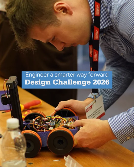 Person assembling a small robotic vehicle with visible wiring and wheels on a wooden table during a design challenge event.