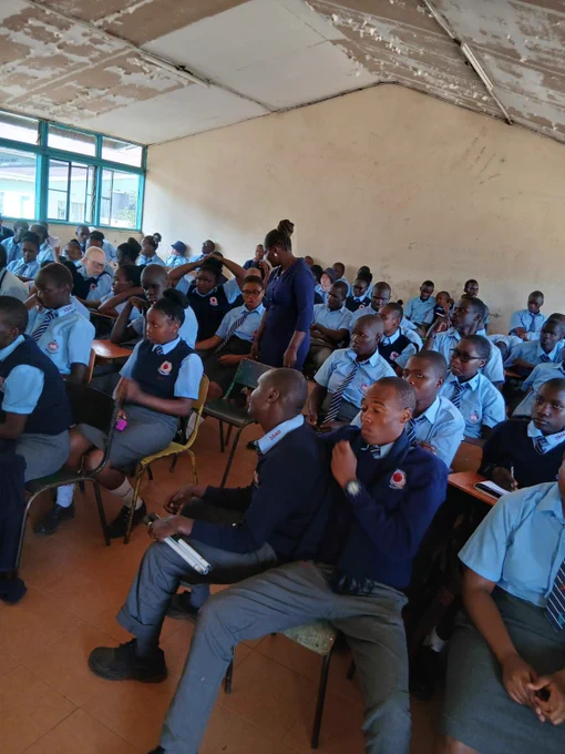 A classroom filled with attentive students in uniform seated at desks, listening to a teacher walking among them.