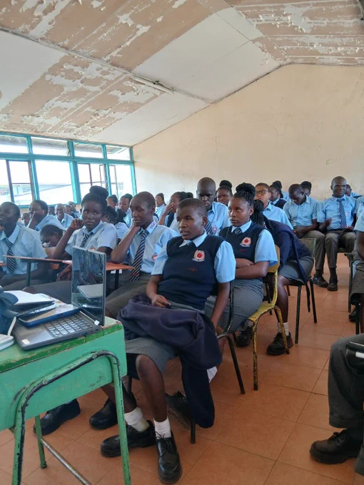 Students in school uniforms attentively seated in a classroom with peeling ceiling paint and large windows letting in natural light.