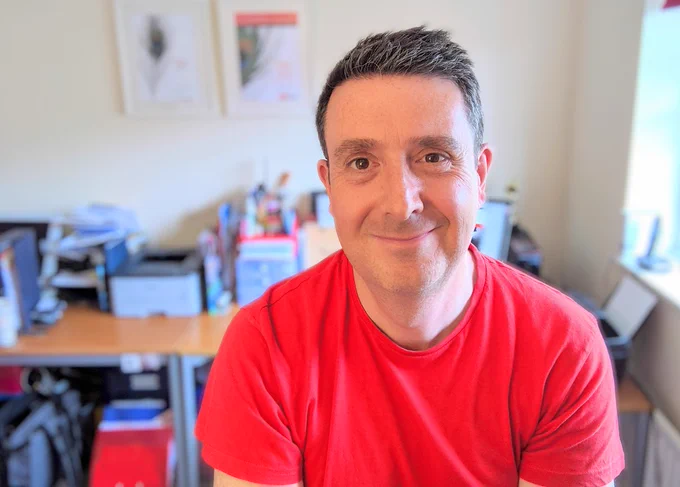 Man in red T-shirt in front of busy office desk.