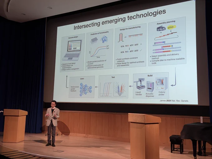 Patrick Cai stands on a university stage in front of a presentation screen describing his work. The slide is on "intersecting emerging technologies" and displays automation equipment including the PIXL colony picker from Singer instruments. 