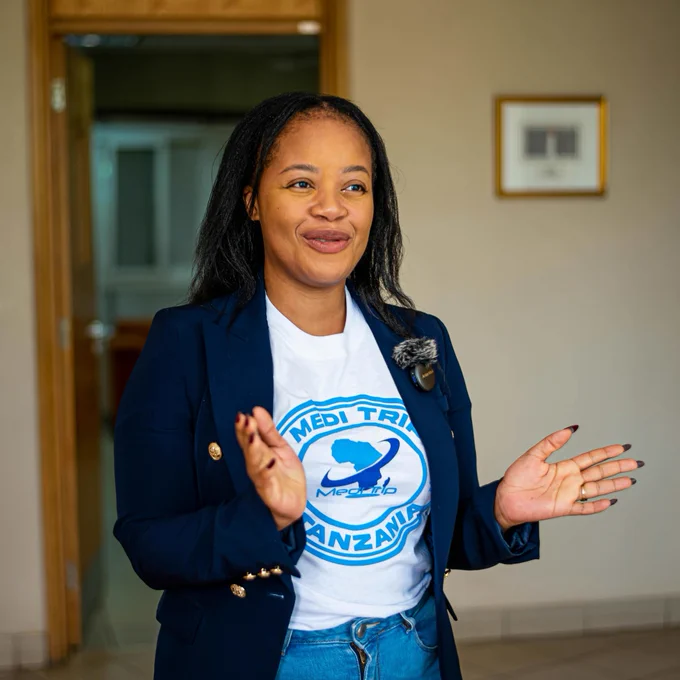 A young woman with long hair in a blue jacket and jeans. Her t-shirt has the words ‘Medi Trip Tanzania’ on it.