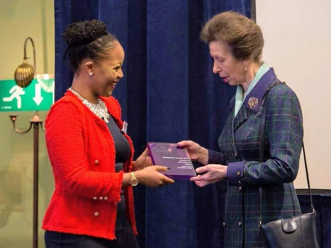 A young woman in a stylish and a dark top receives a plaque from a woman wearing a tartan coat at an awards ceremony.