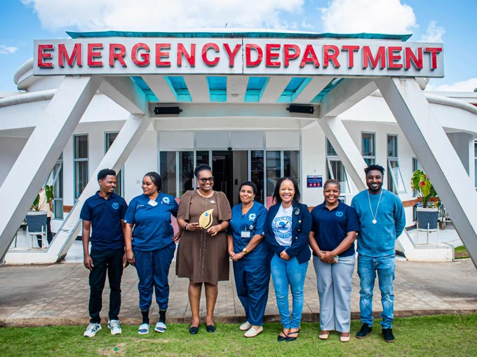 A group of seven men and women stand outside a hospital. The words ‘Emergency Department’ are visible over their heads on a large sign. Five of the group wear medical scrubs and a woman wearing a brown dress holds an award. Also present is a young woman with long hair in a blue jacket and jeans.