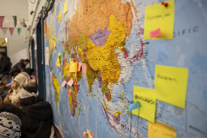 Image of a map in our drop-in centre where people have marked their country of origin, with lots of post-its in East Africa, plus some reading 'Azerbaijan', 'Sri Lanka' and 'Bangladesh'. People sit drinking tea in the background.
