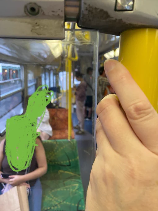 A photo of my hand, holding onto a pole on the tram, with my fingers just fitting into the gap between the pole and the glass panel. In the background are other passengers, and the iconic green and orange fabric on the seats.