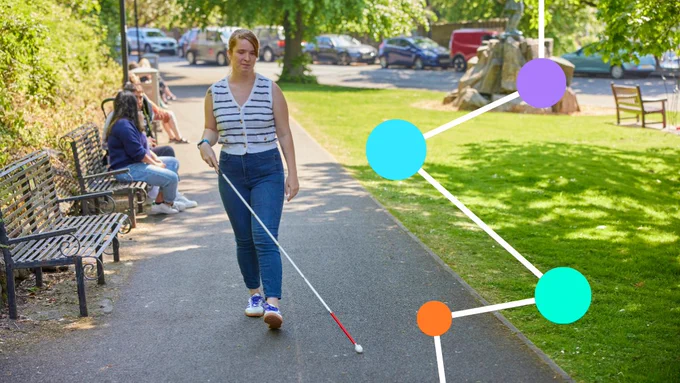 A young women wearing a striped top and blue jeans, and holding a white and red walking aid walks along a path through an urban park. Sunlight shines through the nearby trees.