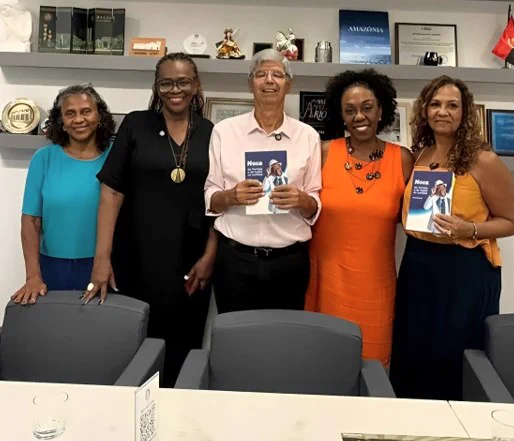 A imagem mostra cinco pessoas em pé, posando para uma fotografia em um ambiente institucional. Entre elas, estão o reitor da UFRJ, Roberto Medronho, que segura um exemplar de um livro do compositor Noca da Portela. Ao seu lado, estão Denise Francisco Góes, da Superintendência-Geral de Ações Afirmativas, Diversidade e Acessibilidade (Sgaada), e Cecília Izidoro, também da Sgaada. Ao lado delas, mais duas mulheres. Na parede, ao fundo, há estantes com diversos objetos decorativos, placas, certificados, prêmios e livros, o que reforça o caráter formal e acadêmico do espaço.
