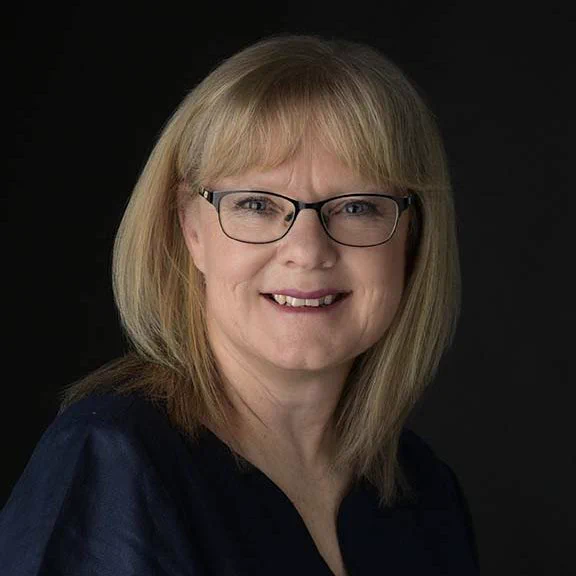 Portrait photo of Bev Smith, the newly appointed Chair of the Local Government Boundary Commission for England. The head-and-shoulders photo shows Bev sitting with her face turned to look at the camera, she is wearing black metal-frame glasses and is smiling.