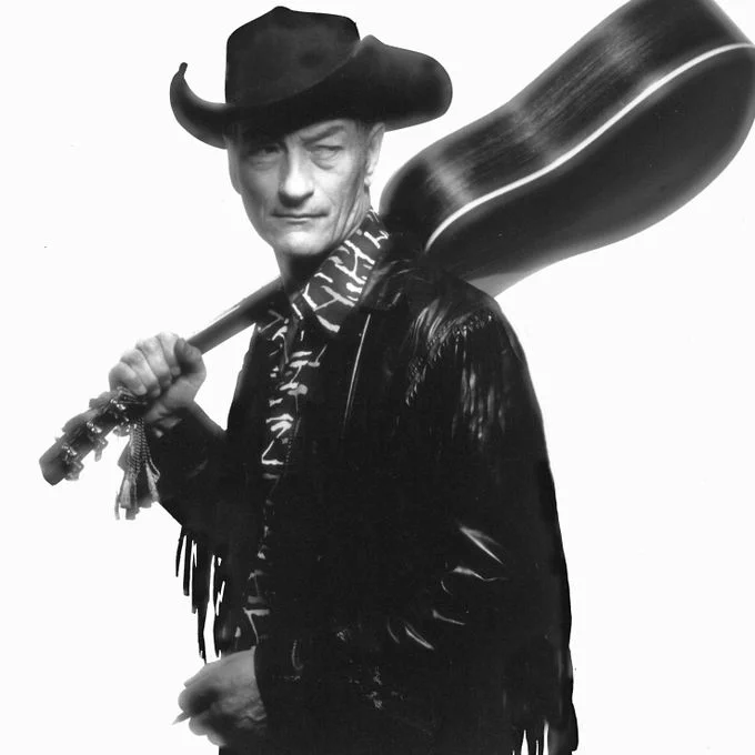 Black-and-white studio portrait of Stompin' Tom Connors standing in three-quarter view against a plain light background. He wears a dark cowboy hat, a fringed jacket, and a patterned scarf. An acoustic guitar in a hard case rests over his right shoulder, held by the neck, with the headstock and tuning pegs visible near his hand. He looks slightly to his left, with one hand near his waist and the other supporting the guitar case.
