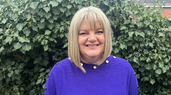 Laura Haslam has a blonde bob and wears a blue jumper. She stands outdoors for a photo, in front of a green hedge.