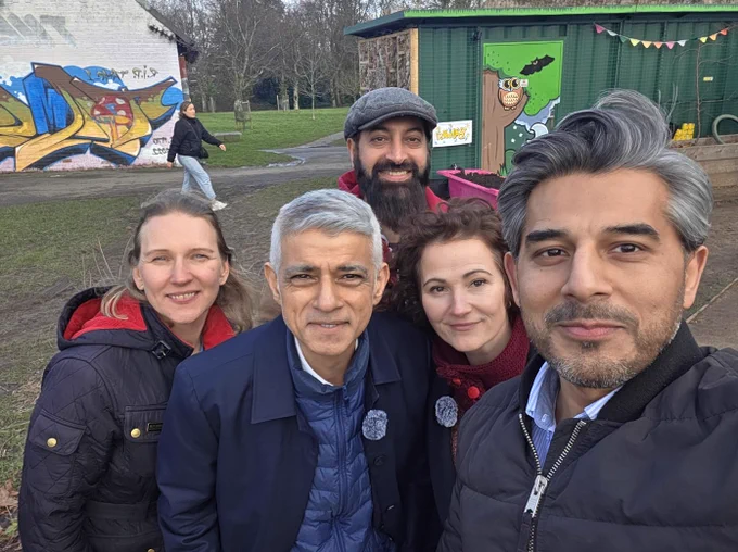 A group selfie taken in Cringle Park, Levenshulme. In the foreground, Cllr Basat Sheikh holds the camera. Standing behind him are Cllr Dzidra Noor, the Mayor of London Sadiq Khan, and Angeliki Stogia, the Labour candidate for the Gorton and Denton by-election. Cllr Zahid Hussain stands at the back. Behind them is the Grounded MCR eco-cafe, a green shipping container with an owl mural and colourful bunting.