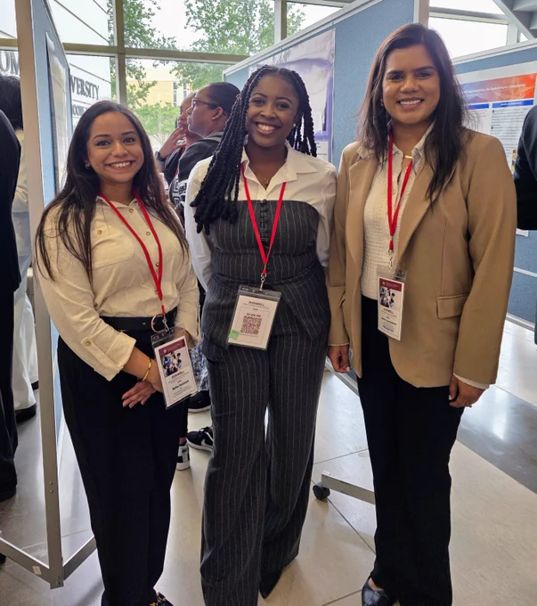 Three attendees smiling at a professional conference, wearing lanyards and standing next to a poster labeled "WESTY.