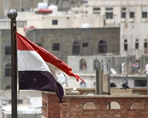 The striped red, white, and black flag of the country of Yemen waves in the wind. Beige buildings stand in the background of the flag. 