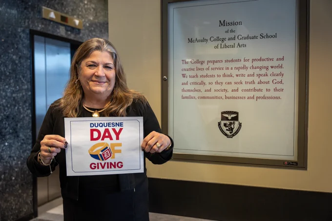 Dean Kristine Blair holds a Day of Giving sign in front of the Liberal Arts mission statement in the second floor College Hall lobby.