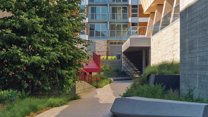 A modern residential building with large windows is seen in the background. In the foreground, there is a winding paved pathway surrounded by lush greenery, a stone retaining wall, and a vivid red staircase.

Photo by Michael Wells Photography