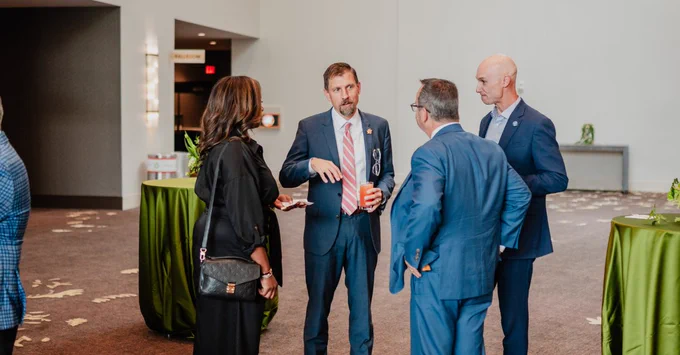 Mason Ailstock, CEO and President of Rowen, engaged in conversation with 3 other attendees at an event. They are standing near tall, round tables with green tablecloths. 