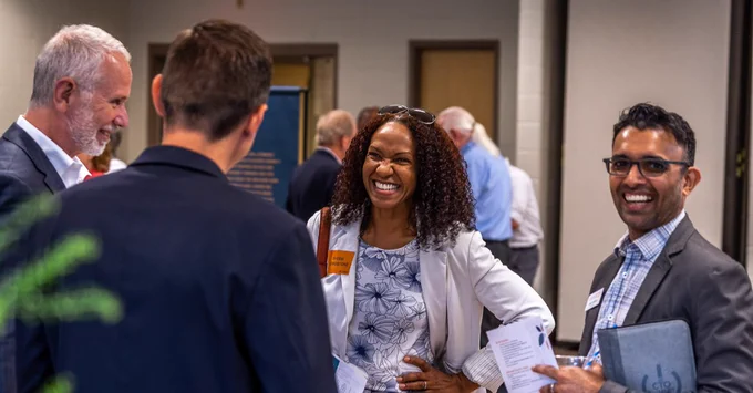 Four people are engaged in a lively conversation at an indoor event. One is holding papers and another is gesturing with a smile. The group appears to be enjoying their discussion. In the background, other attendees are visible, enhancing the sense of a bustling atmosphere.