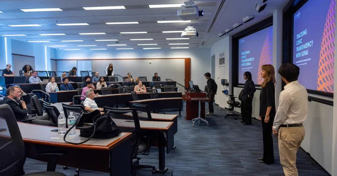 A group of people are sitting in a lecture hall with tiered seating, facing students presenting in front of two large screens at Emory University.