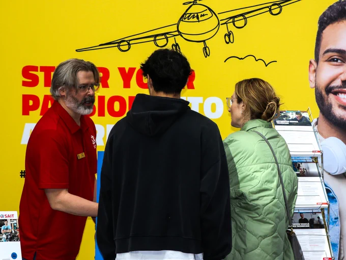 Three people stand near a booth at an event. One person in a red shirt is talking to two others. Behind them, a yellow wall reads, "START YOUR PASSION INTO ACTION" beneath a simple airplane drawing. Brochures are displayed on a stand.