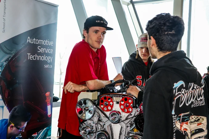 Three individuals are gathered around an engine display. One person is leaning on the engine, wearing a cap and red shirt, while another stands nearby. The third individual wears a dark hoodie with a racing logo. A banner for "Automotive Service Technology" from sait.ca is visible in the background.