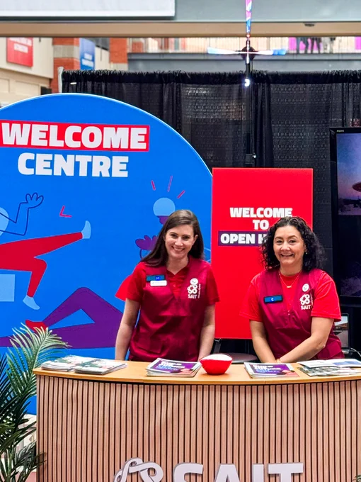 Two individuals wearing SAIT-branded red clothes stand behind the welcome centre. 