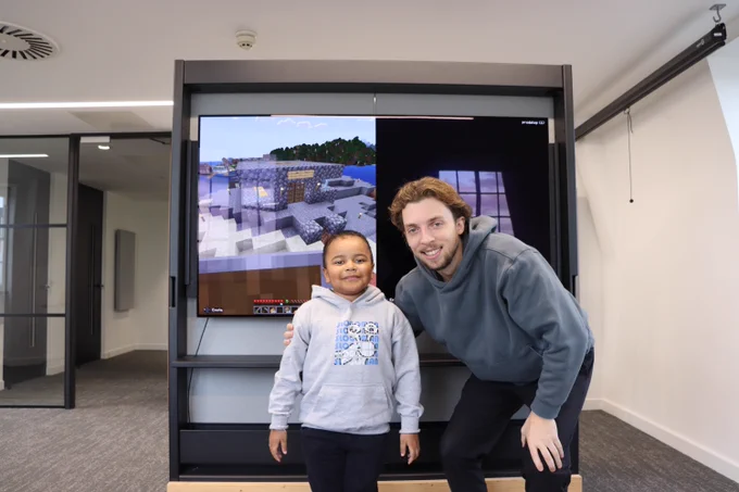 A young boy and a man pose in front of a large screen displaying a Minecraft game. Both are smiling and wearing casual hoodies.