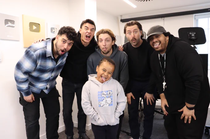 Five adults and one child make funny faces and stick out their tongues in a bright room. Plaques on the wall suggest a social media setting.