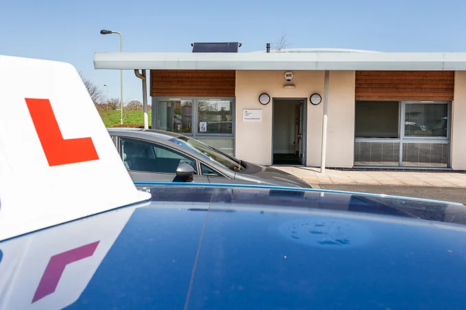 Blue car with a red learner driver 'L' sign parked outside a small single-story building under clear blue sky.