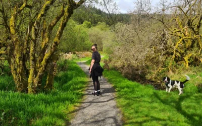 Nikki in black outfit walking on a countryside path with a black and white dog nearby on green grass.