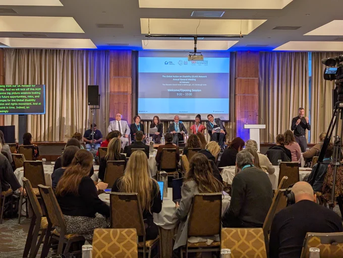 Another image from the back of a conference room with people facing towards a stage with a group of mixed panellists and a large screen behind. 