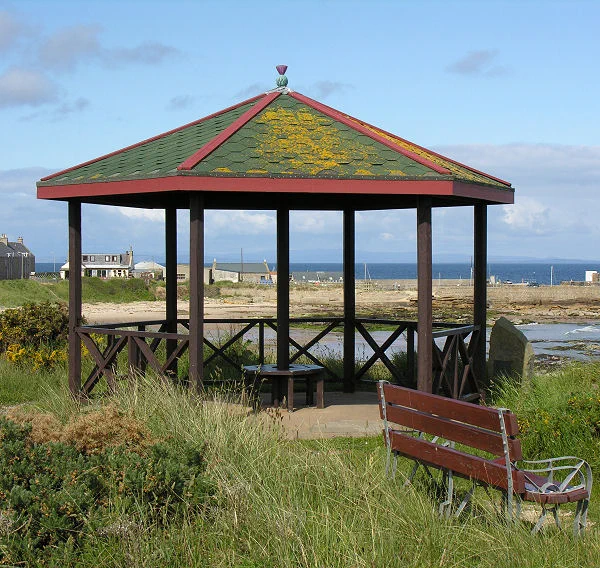 Today's 'Where in Scotland' image. It is a squarish image showing a small hexagonal pavilion with a grey slate roof stained by yellow moss. It is supported by seven dark wood posts and has a low lattice wooden structure between the edge supports and a seat surrounding the central one. There is a bench in the bottom right of the frame and in the background is a coastal scene with sea and sand. The sky is blue.
