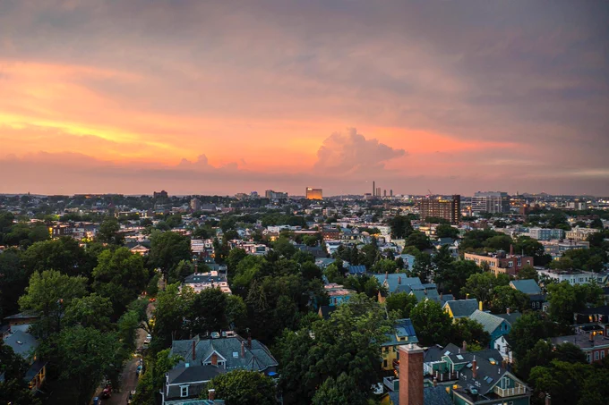 Aerial view of a residential area at sunset with a wide expanse of trees and houses. The sky displays pink and orange hues, and there are tall buildings in the distant horizon.