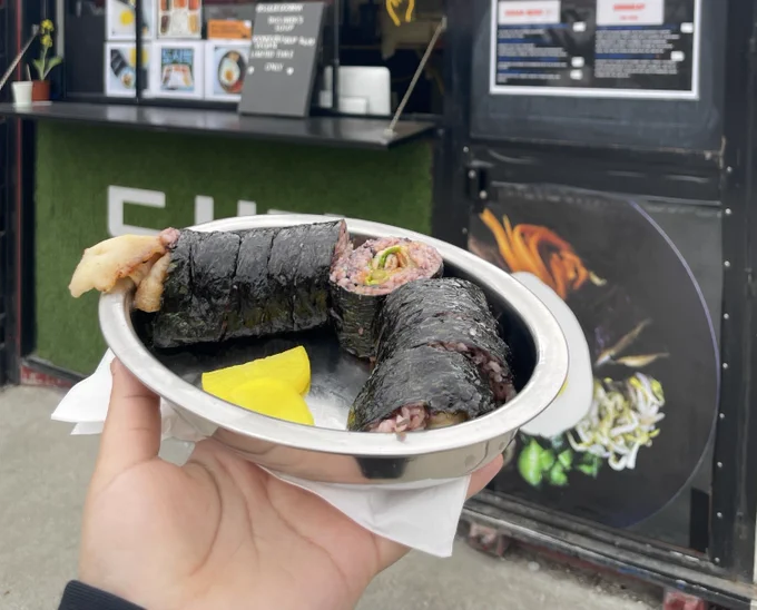 Someone holds an oval silver bowl with a roll of seaweed and rice in front of a food stand.  