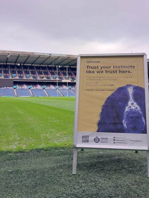 Image shows Murrayfield Stadium in Edinburgh with Project Servator sign. The sign reads 'Trust your instincts like we trust hers. I search for things that could put others in danger and am fearless when confronting threats. Call us on 101. In an emergency always dial 999. Together we've got it covered.' The sign features an image of a cocker spaniel police dog.