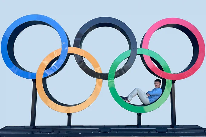 Jerry Lu is sitting inside the green ring of a large Olympic Rings sculpture against a clear blue background.