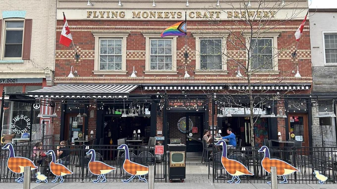 The image shows the front of the Flying Monkeys Craft Brewery. It has a brick façade with a sign displaying the brewery's name. The entrance area is decorated with flags, including Canadian and rainbow flags. A row of geese decorations line the outdoor seating area.