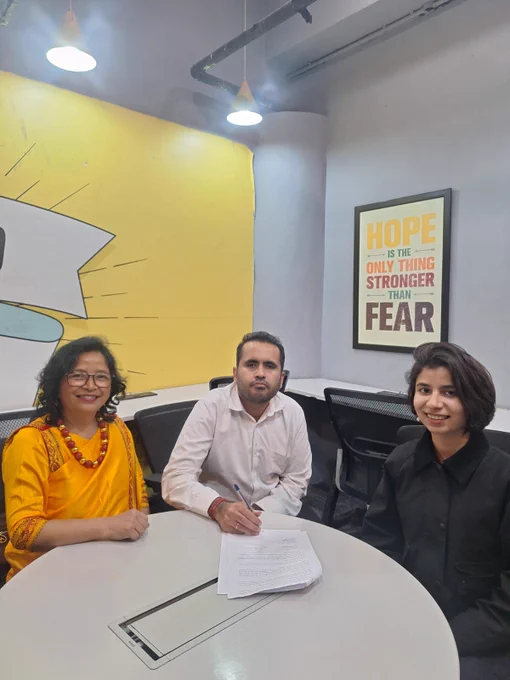 Three people seated at a round meeting table during an MoU signing ceremony at Mission Accessibility’s office. Rahul, Co-Founder of Mission Accessibility, is signing the document. Seated beside him are Sarah and Brigit Warshong, Meghalaya Deputy State Commissioner for Persons with Disabilities. The background features a yellow wall with a graphic design and a framed poster reading “Hope is the only thing stronger than fear.” The setting appears to be a modern office meeting room.